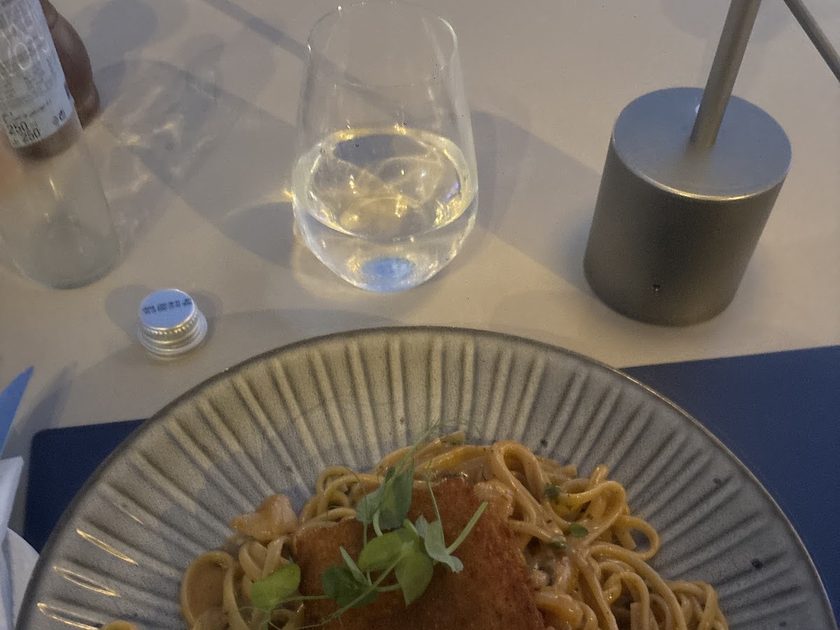 Creamy linguine topped with a breaded fillet and pea shoots, beside a glass of water
